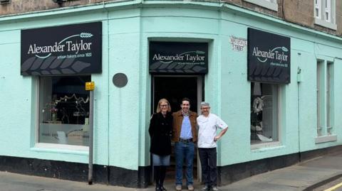 Barry and Claire Taylor with Carlos Huerta standing is front of Alexander Taylor bakery. The bakery is a blue-green painted building on the corner of a street with signs reading Alexander Taylor above the doorway and windows. Claire is wearing a black coat and blue jeans, Carlos is wearing a blue shirt and jeans with a brown jacket, and Barry is in chef whites.