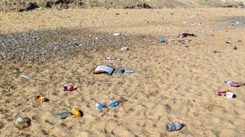 Litter including cans, cardboard packaging and bottles discarded on a sandy beach on a sunny day.