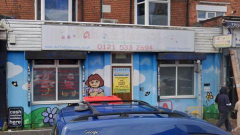 The outside of Daffodils Nursery in Smethwick. The walls are painted in a mural showing blue sky and flowers and a character standing by the door. There is a faded sign above the entrance in Waterloo Road.