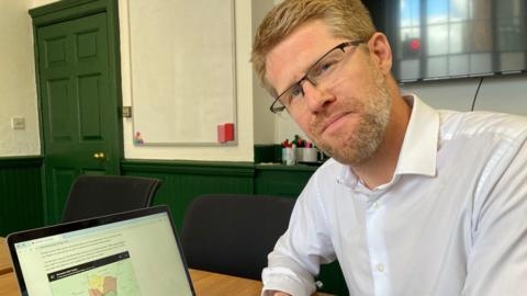 A man sat at a desk is wearing a white shirt. In front of him is a laptop, which shows a map on it. 