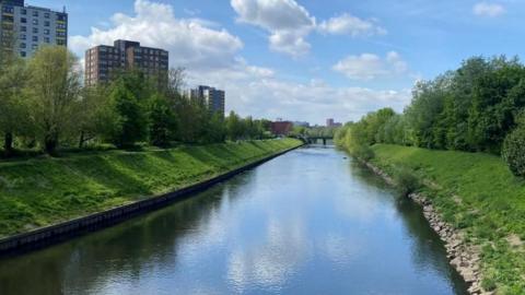 A river is surrounded by green grass and trees as well as apartment buildings. 