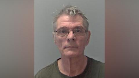 A police custody image of a man with grey hair looking directly at the camera. He wears glasses and a green T-shirt.