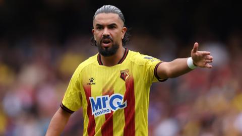 Watford's Imran Louza with his arm out. He is wearing a yellow and red striped kit. 