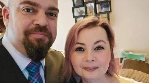 Laura, who has pinkish shoulder-length hair, stands next to her husband, who is wearing a colourful tie and jacket 