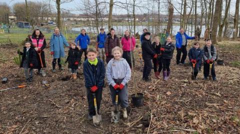 School children in Killingworth Lakeside Park during a visit to plant trees. A boy and girl are standing at the front of the group and are each holding a spade. A potted tree stands next to them. A group of children and teachers stands behind the. In the background are existing trees, a lake and a play park.