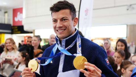 A man holding two Olympic medals. He is wearing a Team GB blue fleece. Around ten people look on in the background.