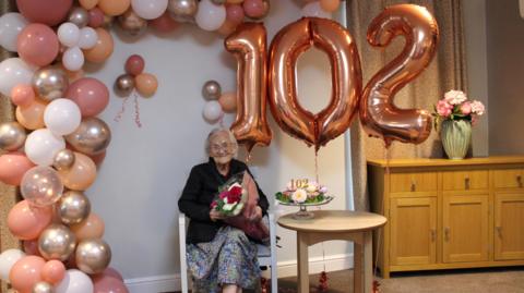 An elderly woman sits in a white chair. Balloons in shades of pink, white, gold and orange surround her and there is a balloon in the shape of the numbers 102 next to her. A small circular wooden table rests next to her where there is also a cake with a 102 on it.