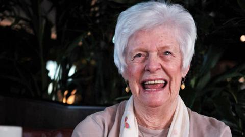 Marie McLaren smiles for the camera. She has short grey hair and is wearing a white scarf.