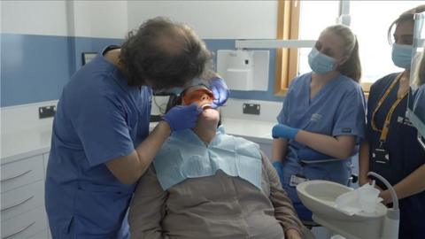 A dental patient lies in a chair as a dentist works on her teeth. Two other dental staff who all wear masks watch on.