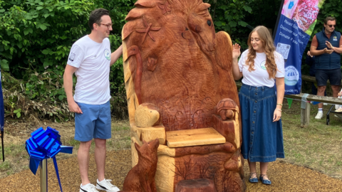 A large wooden throne, with animals including a horse, a fox and a hedgehog carved into the chair. Two people in white t-shirts are stood either side of the installation.