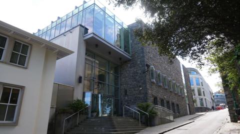 Guernsey's Royal Court - Modern multi-storey building with a stone and glass façade situated on a sloping street. The entrance features wide steps, metal handrails, and a revolving glass door beneath a large glass-fronted upper level. Trees overhang the roadway on the right, while additional office-style buildings line the street in the background. 