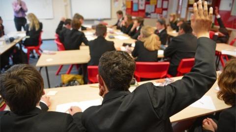 The backs of teenagers in a classroom with a boy raising his right hand