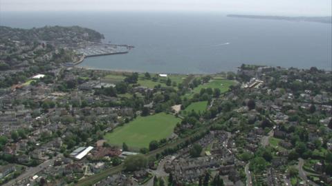 A drone shot from high in the sky overlooking Torquay, Devon. There is lots of buildings and green spaces as well as the sea in the distance.