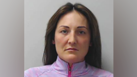 Alisha Bain, a woman with long dark hair and wearing a pink/purple zip top, staring at the camera, unsmiling, in a police mugshot.