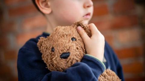 A small boy in a blue jumper is clutching a brown teddy bear