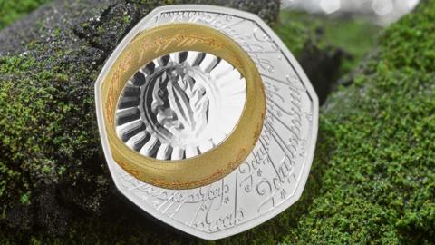 A single coin on a fake background of granite grey rock frosted in green lichen. the coin is silver with an etched inscription in Elfish, a Lord of the Rings script and language. A gold band, the one ring, also inscribed in Elvish, is at the top left of the seven sided coin. the ring surrounds the eye of sauron etched onto the silver coin with an oval around the eye which is surrounded by a fan of lines to the ring. 