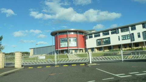 A modern school building with a round red section and the rest in white behind gates