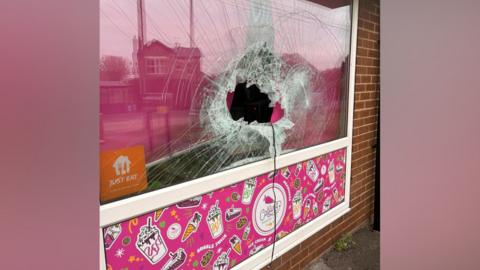 Catling Bakery's smashed window. A large hole has been made in the middle of the pane of glass and cracks have spread across the rest of the window. A pink banner below the window shows cartoons of cakes and milkshakes.