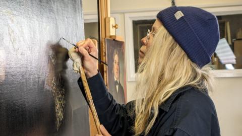 A woman with long blonde hair touches up a dark portrait on an easel with a very thin paintbrush. She wears glasses, a blue beanie hat and a dark blue shirt.