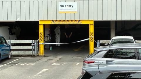 A person in white overalls can be seen beyond a yellow entrance into a car park, with police tape blocking the entrance. Parked cars are visible in the foreground
