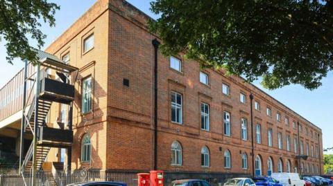 A picture of the outside of the building. It is a large brick building with cars parked at the front of it.