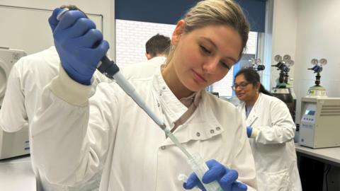 A woman in a white medical coat holding a pipette and a sample pot