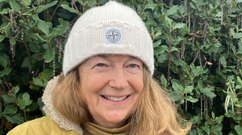 Person wearing white woollen hat and a yellow coat stands in front of green foliage