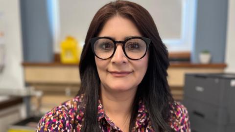 Dr Qayyum has long dark hair and is smiling at the camera. She is wearing black round framed glasses and a purple, pink and red patterned blouse. 
