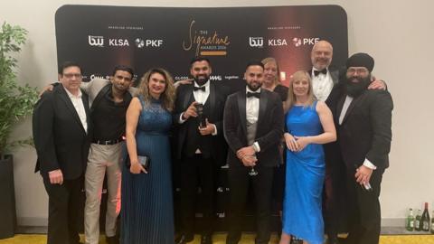 Nine people are standing together in front of a black banner with gold writing on it which reads "the signature awards 2026". There are six men and one woman. The men are dresses in suits and ties, and the women are in dresses. In the centre is Manny Athwal, who is in a black suit with a white shirt and black dickie bow tie. He is holding a black trophy.
