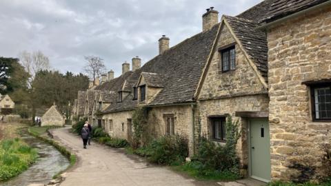 A row of Cotswold stone cottages in Bibury. A man and a woman are walking along a path nearby. A stream runs on the left hand side of the path.