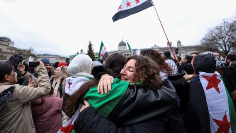 Two women hugging at a demonstration in Trafalgar Square. One has the flag of Syria draped on her back. There are surrounded by other people who also have the Syrian flag around their backs while some are holding flags.