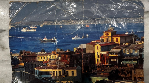 A dog-eared post card depicting a coastal community, with boats in a bay flanked by old, multi-storey homes.