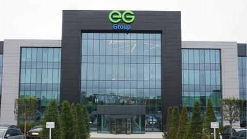 The EG group headquarters, a large building with a rectangular grey frame and lots of glass windows. In the middle is a taller black framed structure, also with many windows and the EG group logo in green and blue at the top