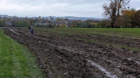 Muddy ground in South Park, Oxford, showing vehicle tracks through the grass after the 5 November 2023 fireworks display. A couple of people could be seen in the middle of the muddy tracks far away from the camera. The Oxford skyline can be seen in the distance