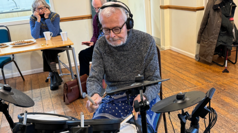 Nick Foster is sitting at an electric drum kit. He is wearing a grey jumper and has headphones on.