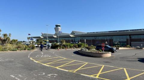 Jersey Airport on a sunny and clear blue sky day. There is a man walking up to the airport building which is largely glass with three triangular shaped glass roofs. The air traffic control tower, a silver spherical building, can be seen just past the airport. 