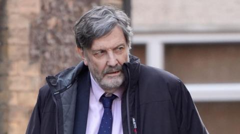 Alan Harris walking. He is wearing a pink top with a blue tie and blazer. He is wearing a black coat. The background shows a brown wall