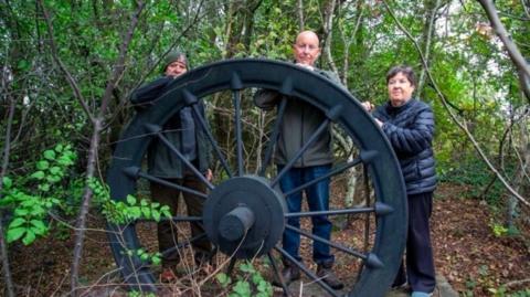 Three people stand behind a large iron mining wheel surrounded by small trees.