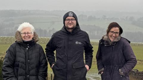 Three people, Kate Culverhouse of the Banks Group, Andrew Wadds of Durham Wildlife Trust and Louise Harrison of the Banks Group, standing in a line in front of a field. It is a grey day and the hills behind them are covered in mist. Kate has white, short hair and is smiling into the camera with one eye closed. She is wearing a big, dark coat and has round glasses on. Andrew is squinting at the camera and is wearing a beanie. He has his hands in his pockets and has a coat on with the Durham Wildlife Trust logo. Louise has dark hair and is wearing glasses. She is wearing a dark coat with her hands in her pocket and is smiling.