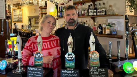 Millie and her husband Jon Coward standing behind a bar looking at the camera. Jon looks serious. Millie has a partial smile.