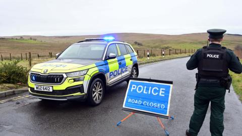 A road with green hills in the background. A police car is parked up beside a blue sign with 'POLICE ROAD CLOSED' written on it. Beside this is a PSNI officer with his back towards the camera. 
