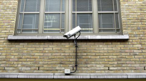 A picture of the side of a brick building. There is a white CCTV camera attached to it in front of a large window.
