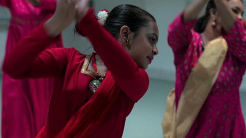 Women in traditional Indian dress practising classical dance movements with raised arms and heads turned to the side