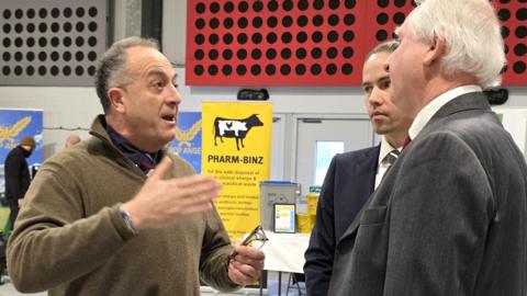 Mark Means is challenging Daniel Zeichner. Mr Means is gesticulating with his hand as he speaks - he's wearing a brown jumper on top of a blue shirt and tie. We can't see Zeichner's face from this angle - but he's wearing a grey suit jacket, white shirt and red tie.