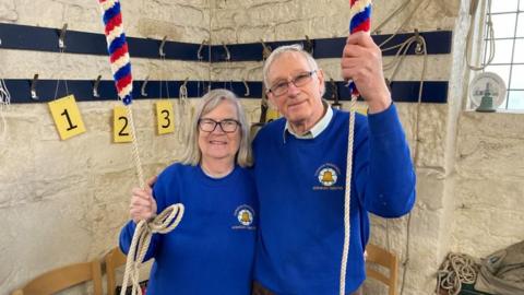 A woman with shoulder-length white hair and glasses and a man with short white hair and glasses hold bell ringing ropes. They are both wearing blue sweatshirts with a gold bell and a white rose on them.