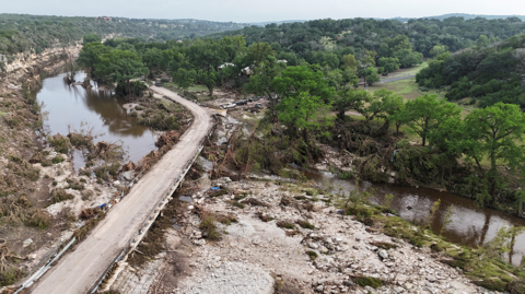 Downed trees and other debris are piled up near a small bridge over a river.