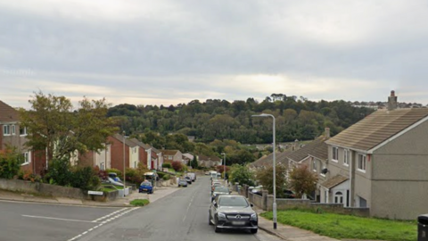 A residential road. Terraced houses line both sides of the street. Cars are parked on the road in front of the houses. There are trees in the distance. A green patch of grass is in front of one house at the end of the street.