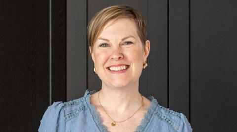 A woman with a brown and blonde pixie cut smiles at the camera. She is in a long-sleeved, light blue V-neck blouse with a lace ruffle around the neckline. The background is black cupboard doors.