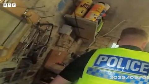 A male police officer wearing a yellow police vest in a shed facing away from the camera