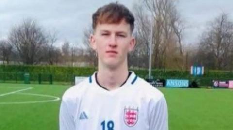 Sam Harding has short brown hair and looks at the camera. He is wearing his England football kit while standing on a football pitch.